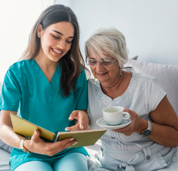 A nurse and an elderly person sitting on a couch reading a book.