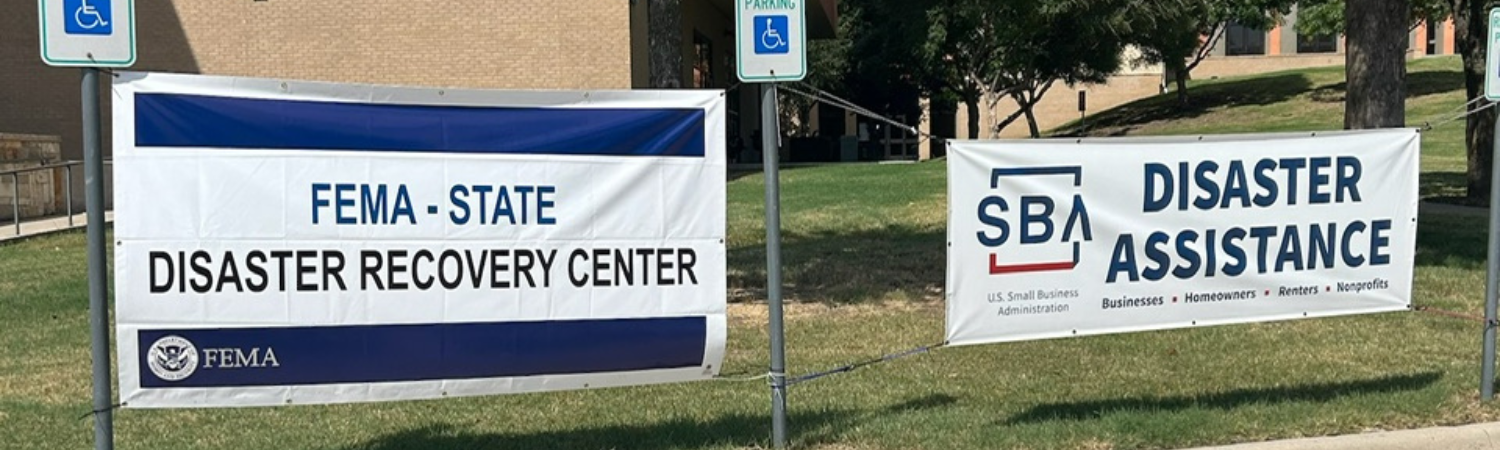 Two signs in front of a parking lot with the words FEMA State Disaster Recovery Center and SBA Disaster Assistance on them.