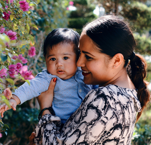 Smiling mother holding her cute baby boy near blooming flowers.